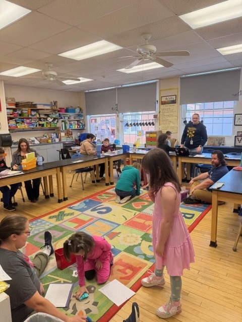 Students and family members spread out across a classroom, working on activities at tables and on a rug, with a teacher standing nearby and classroom supplies lining the walls.