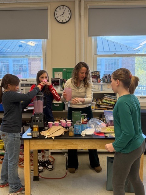 Students and an adult gather around a classroom table using a blender to prepare a drink, with ingredients and cups spread out on the table.