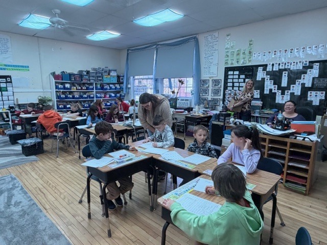 A teacher leans over to assist students working at desks in a bright classroom while other children focus on assignments around the room.
