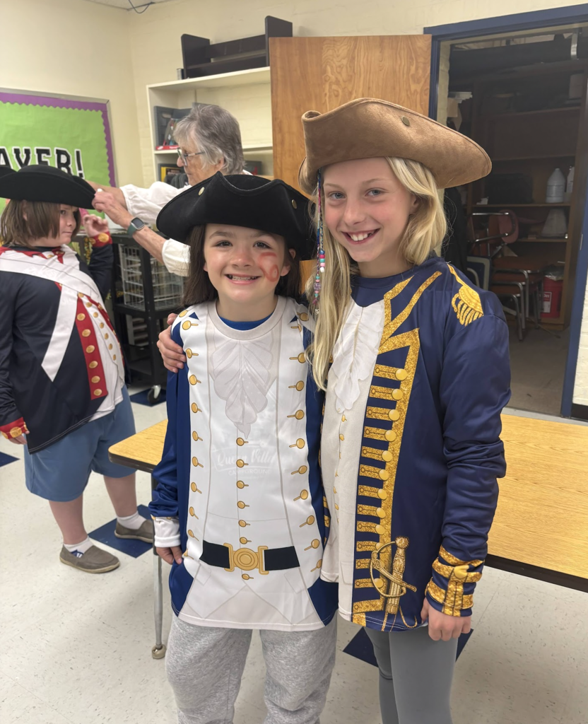 Two smiling students wearing colonial-style costumes and tricorn hats stand side by side, posing for a photo in a classroom.