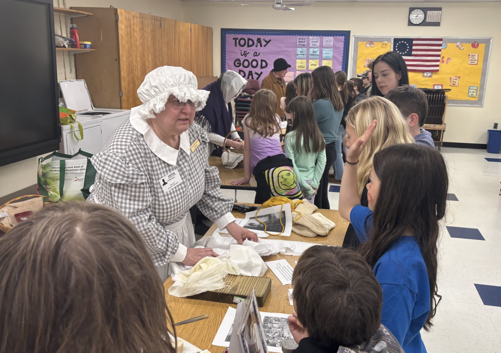 Students stand around a table listening to an adult in historical dress explain materials and artifacts during a classroom activity.