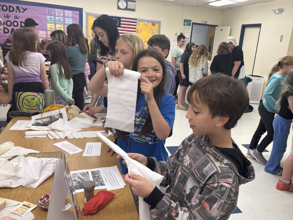 A group of students examine cloth materials at a classroom table while one student holds up a piece of fabric, with others engaged in the activity behind them.
