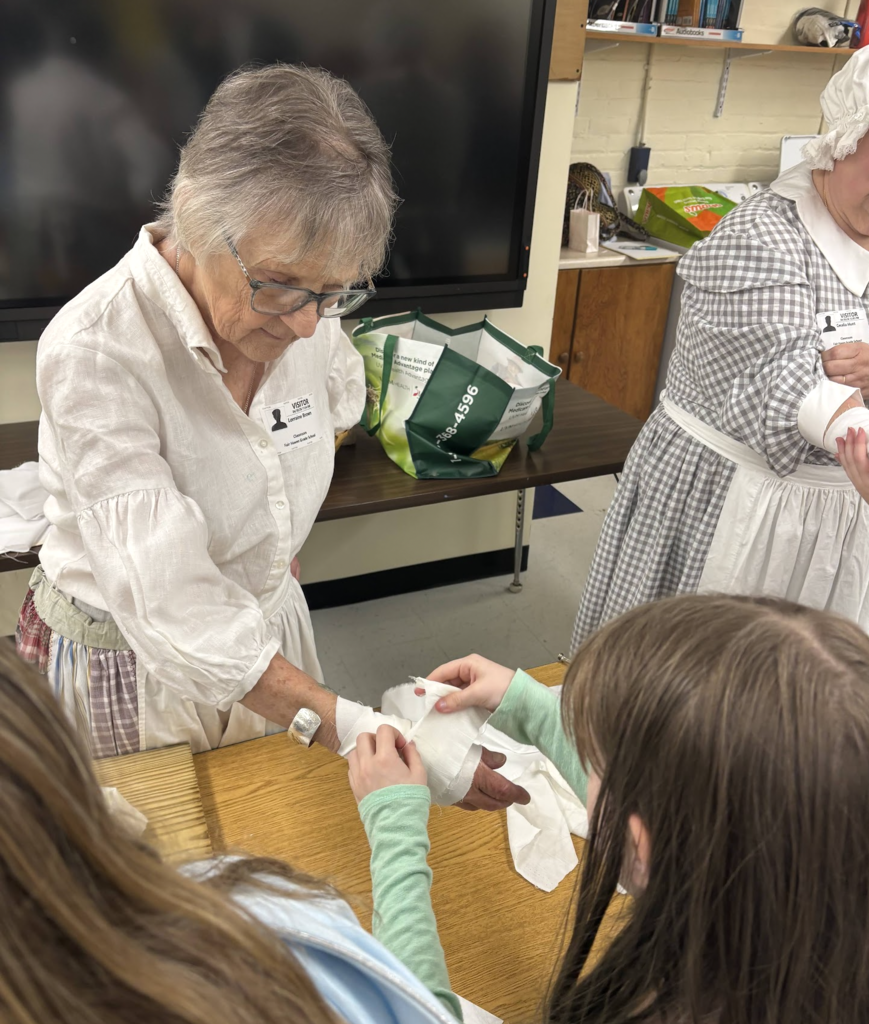 An older adult in historical clothing demonstrates how to wrap a bandage on a student’s arm while other students watch closely around a classroom table.