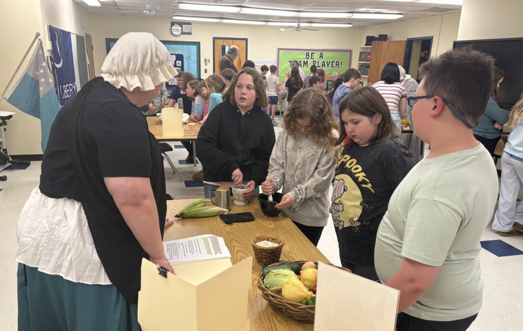 Students gather around a table with an adult in historical clothing, watching a demonstration involving food items like corn and vegetables as part of a hands-on activity.