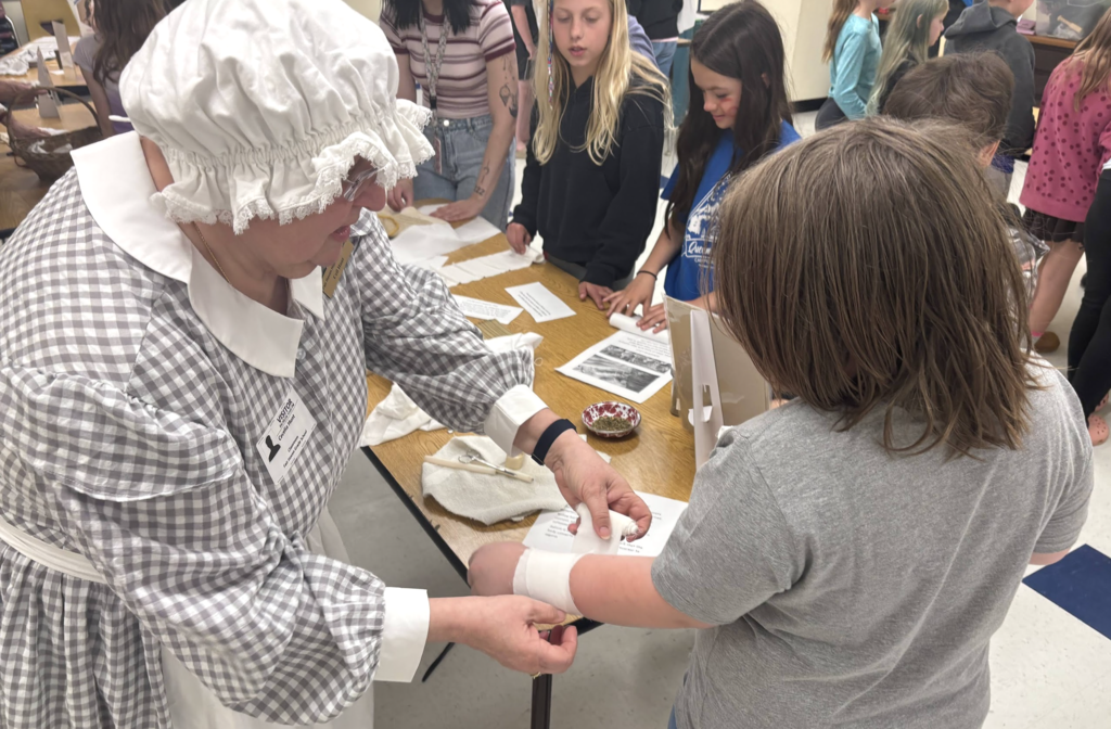 An adult in historical attire helps a student wrap a bandage on their arm while other students observe and materials are spread across a table.