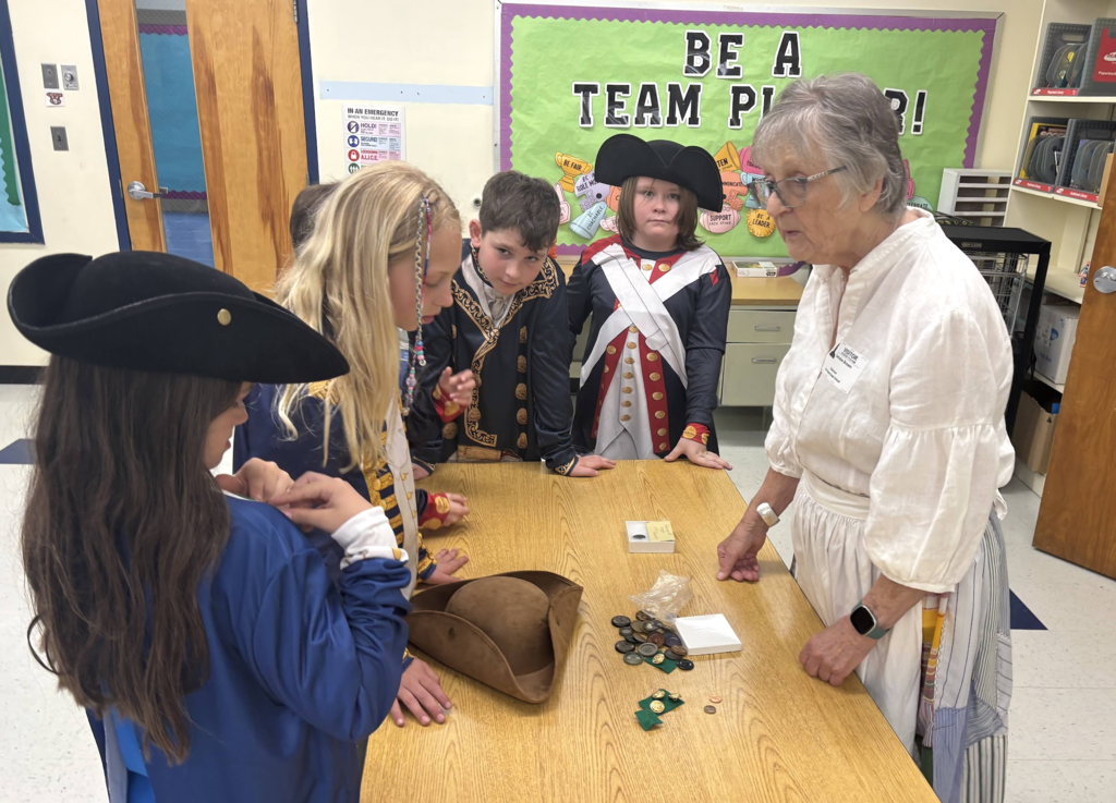 Students dressed in colonial-era costumes gather around a table as an older adult explains historical items, including coins and a tricorn hat.
