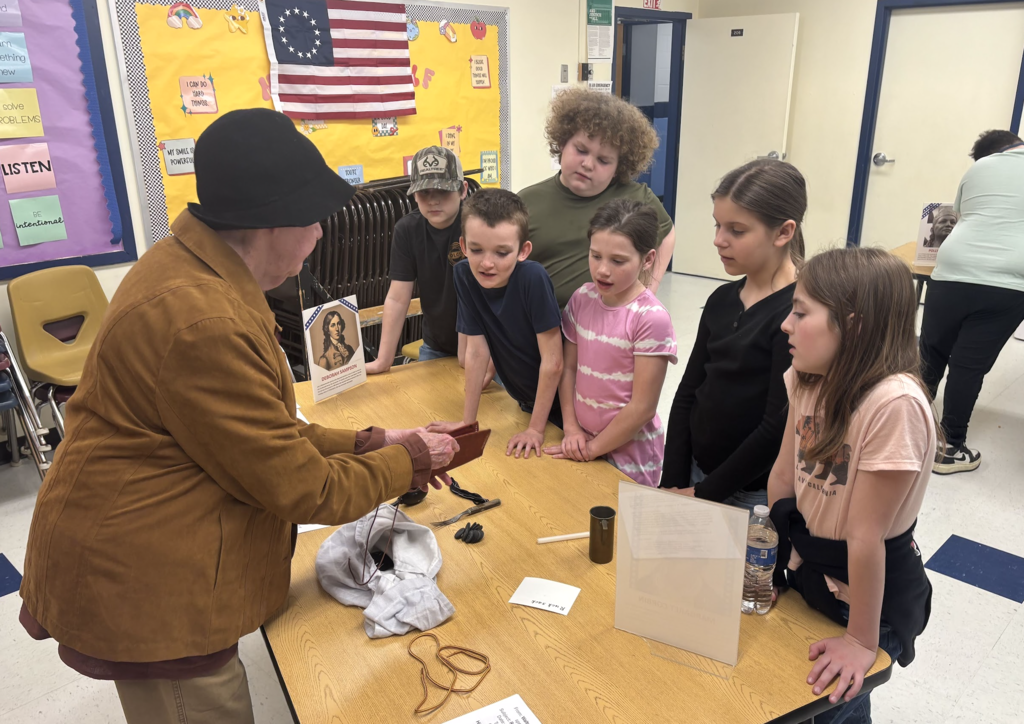 An adult in colonial-style clothing demonstrates an object to a group of students gathered around a table, who watch attentively.