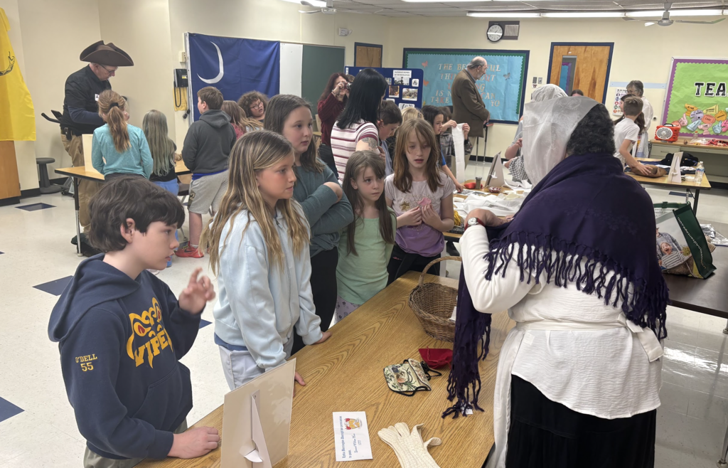 Students stand around a table listening to an adult in historical dress explain materials and artifacts during a classroom activity.