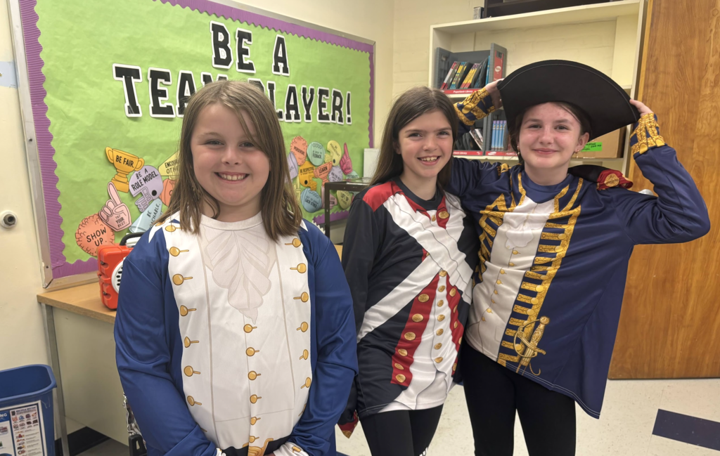 Three students in colonial-style costumes smile and pose together in front of a classroom bulletin board that reads “Be a Team Player!”