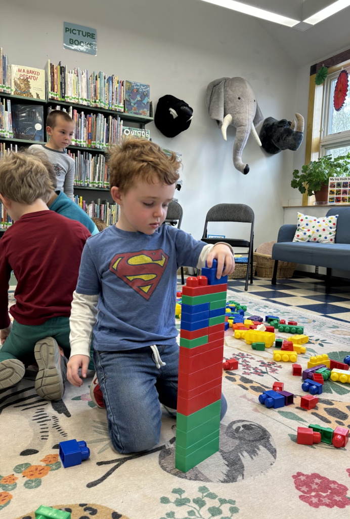 A young student kneels on a library rug, carefully stacking large colorful blocks into a tall tower. Other children and bookshelves are visible in the background.