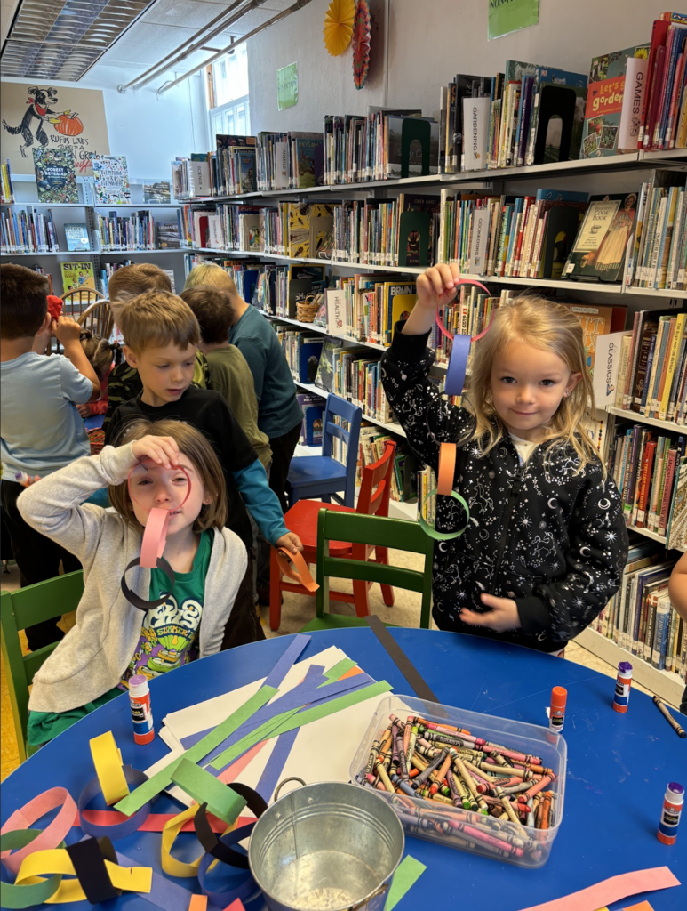 Two young students at a table proudly hold up colorful paper chain crafts they created, while classmates work together in the background of the library.