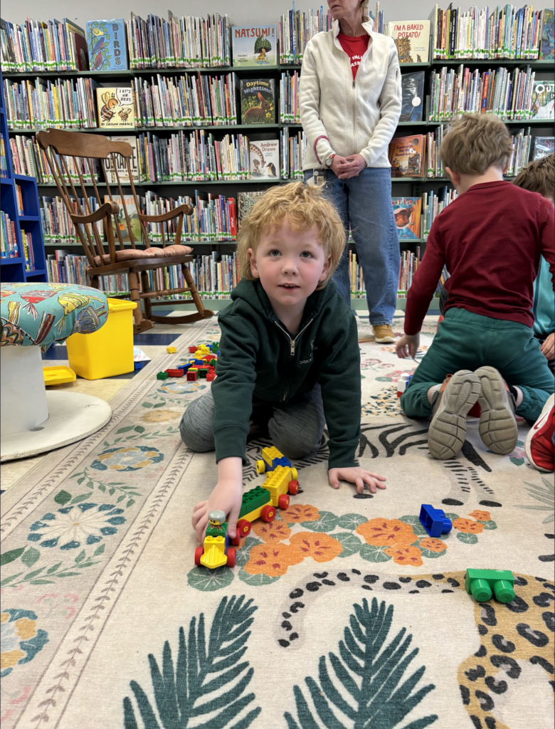 A young student sits on a patterned rug in the library, pushing a small wheeled structure made from large building blocks, while other children play nearby and an adult supervises.
