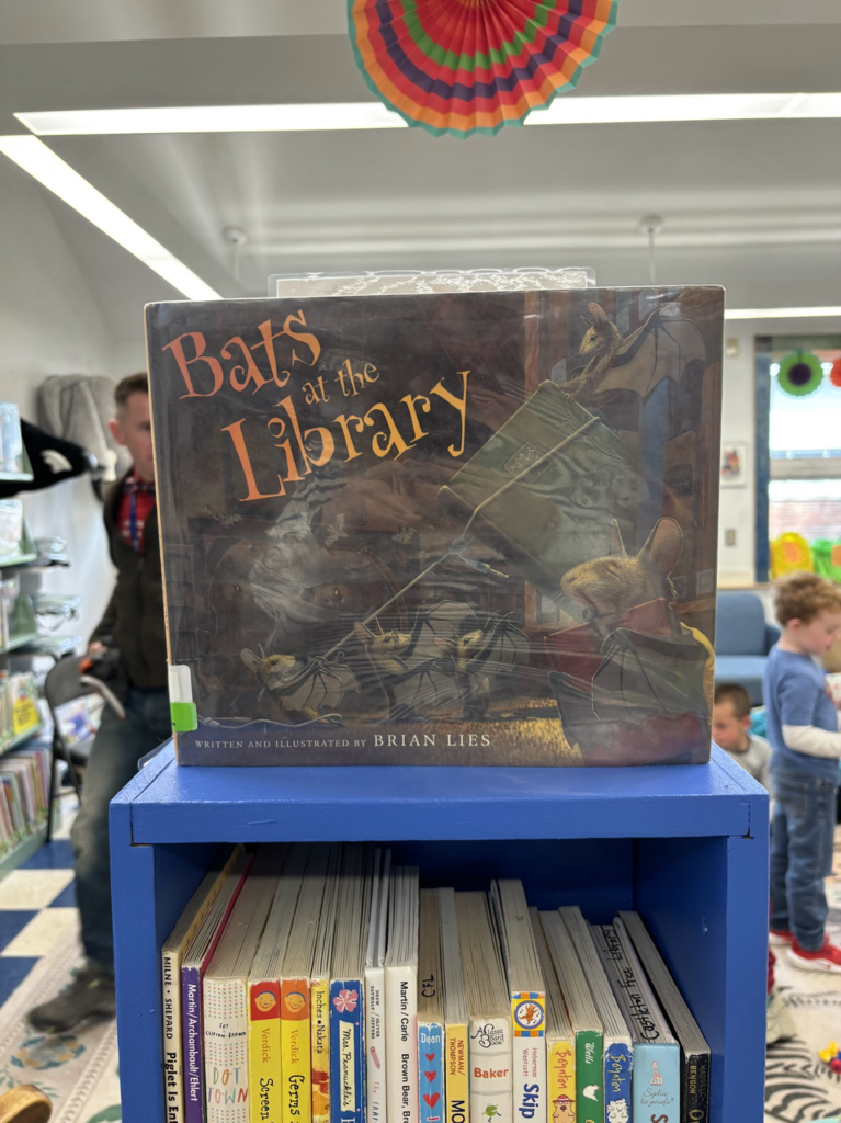 A close-up of the book Bats at the Library displayed on a shelf, with the library space and students blurred in the background.