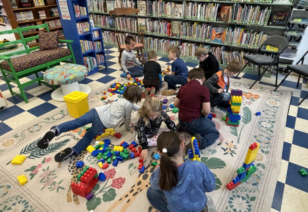 A group of young students gathers on a rug in the library, working together with large building blocks. Chairs, bookshelves, and reading displays surround the space.