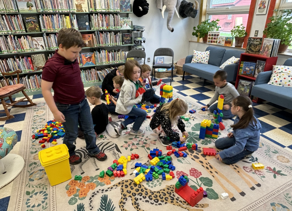 Students sit and kneel on a colorful rug in a school library, building with large plastic blocks. Bookshelves line the walls, and a cozy reading area with chairs and plants is visible in the background.
