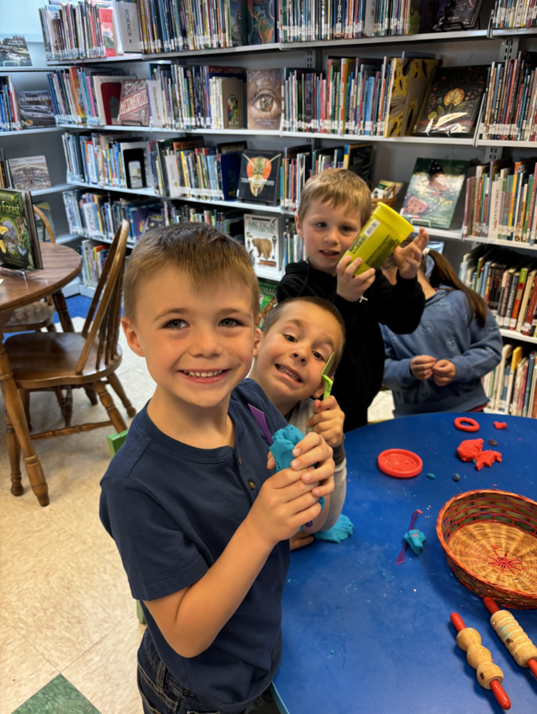 Three smiling children stand at a table in the library, holding modeling clay and tools while showing their creations to the camera. Bookshelves and activity materials are visible behind them.