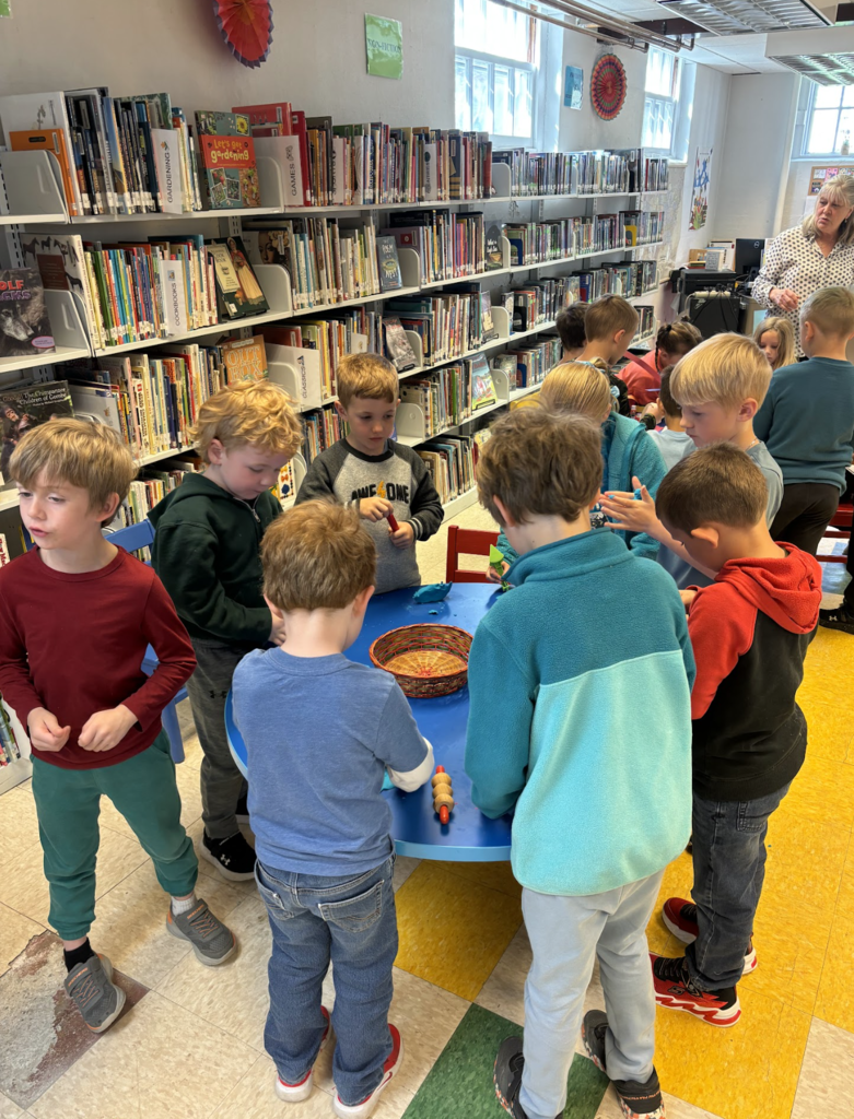Students stand around a small blue table in the library, using hands-on materials to explore and create. Bookshelves filled with children’s books stretch along the wall behind them.