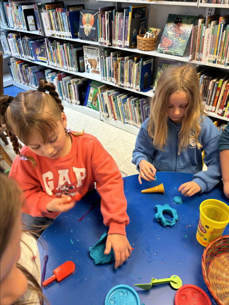 Two students sit at a table in the library, shaping and playing with modeling clay using small tools, surrounded by art supplies and bookshelves.