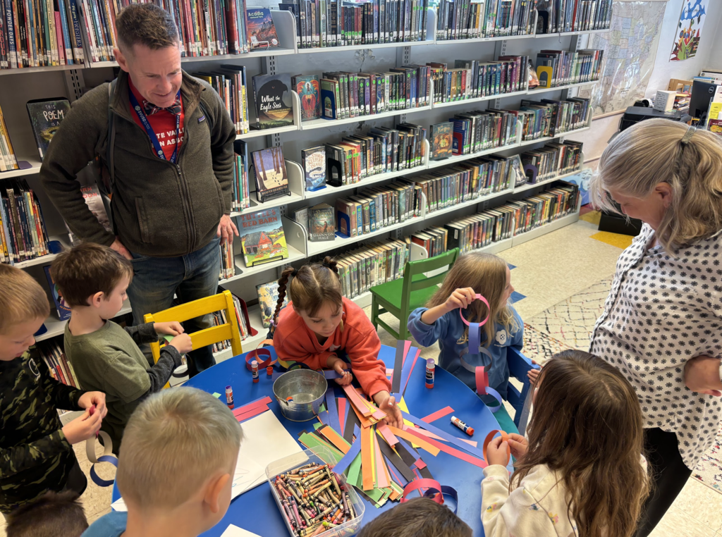 Children and two adults sit and stand around a table in the library, creating colorful paper chains and crafts using strips of paper, glue sticks, and crayons.