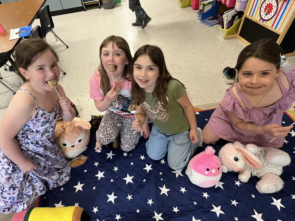 A group of children sitting in a circle on a large blue blanket with white stars. They are in a classroom setting, and many are holding plush stuffed animals (Squishmallows) or small bags of snacks while smiling for the camera.  S’mores Treats (Individuals and Small Groups)