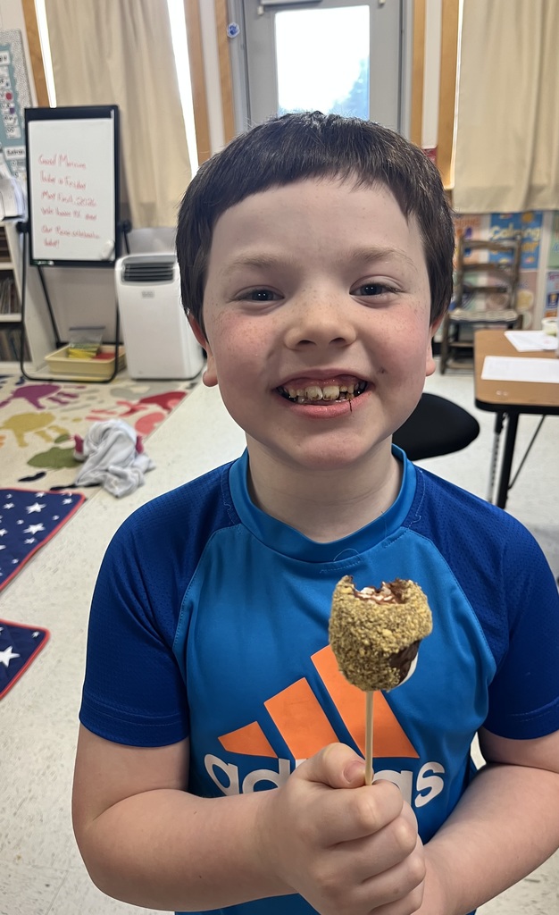 A young boy in a blue Adidas shirt smiling widely while holding a s'more on a stick. He has a bit of chocolate on his teeth from taking a bite.  IMG_5633.jpg: A close-up of a young boy in a white t-shirt grinning enthusiastically as he holds up a marshmallow treat coated in graham cracker crumbs on a wooden skewer.  I