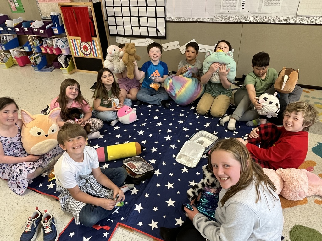  A group of children sitting in a circle on a large blue blanket with white stars. They are in a classroom setting, and many are holding plush stuffed animals (Squishmallows) or small bags of snacks while smiling for the camera.  S’mores Treats (Individuals and Small Groups) IMG_5632.jpg: A young boy in a blue Adidas shirt smiling widely while holding a s'more on a stick. He has a bit of chocolate on his teeth from taking a bite.  