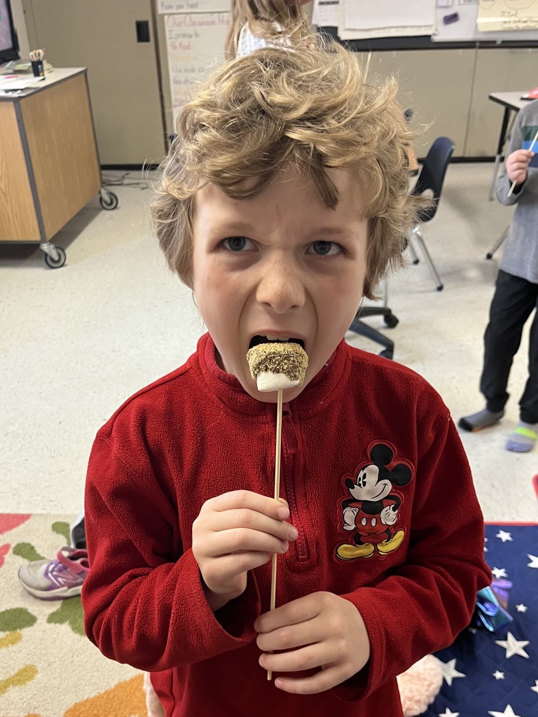 A young boy with curly blonde hair wearing a red Mickey Mouse fleece, opening his mouth wide to take a bite of his s'more treat.  