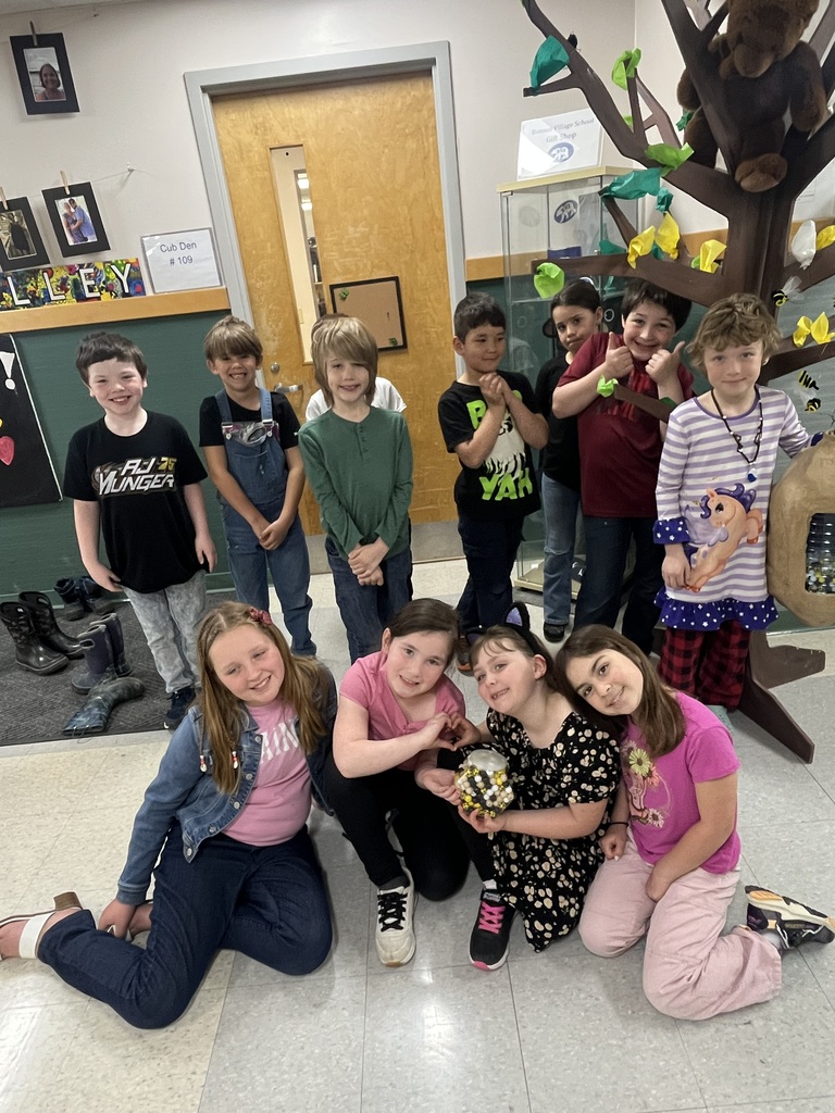 A group of eleven elementary-age children posing together in a classroom. Seven children stand in the back row near a decorative indoor "tree," while four children sit on the floor in front. One of the seated girls holds a small jar filled with yellow and black marbles