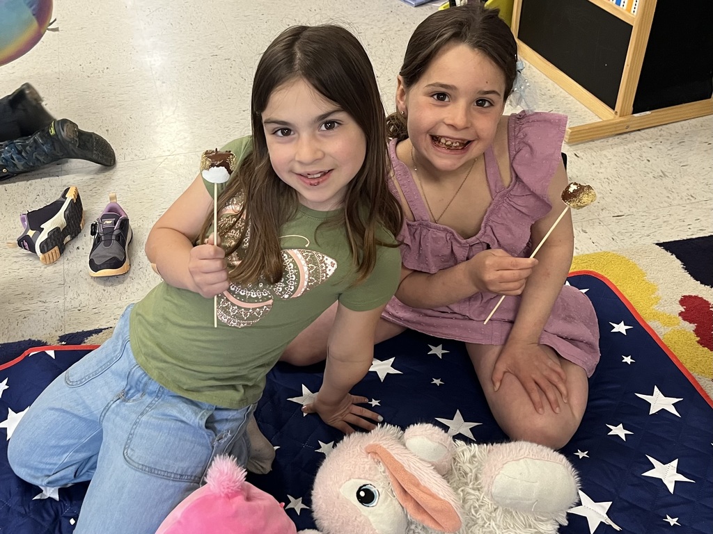 Two young girls sitting on a star-patterned blanket, each holding a s'more on a stick and smiling. A plush bunny sits on the floor in front of them.  IMG_5636.jpg: Four young girls sitting on a blue star-patterned blanket. Two of the girls are mid-bite, eating their s'more treats, while the other two smile at the camera. 