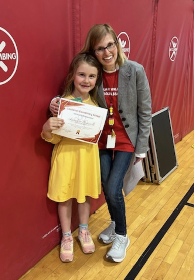A smiling adult staff member kneels beside a young student in a yellow dress who is holding a Castleton Elementary School certificate. They pose together in a gym with red wall padding behind them.