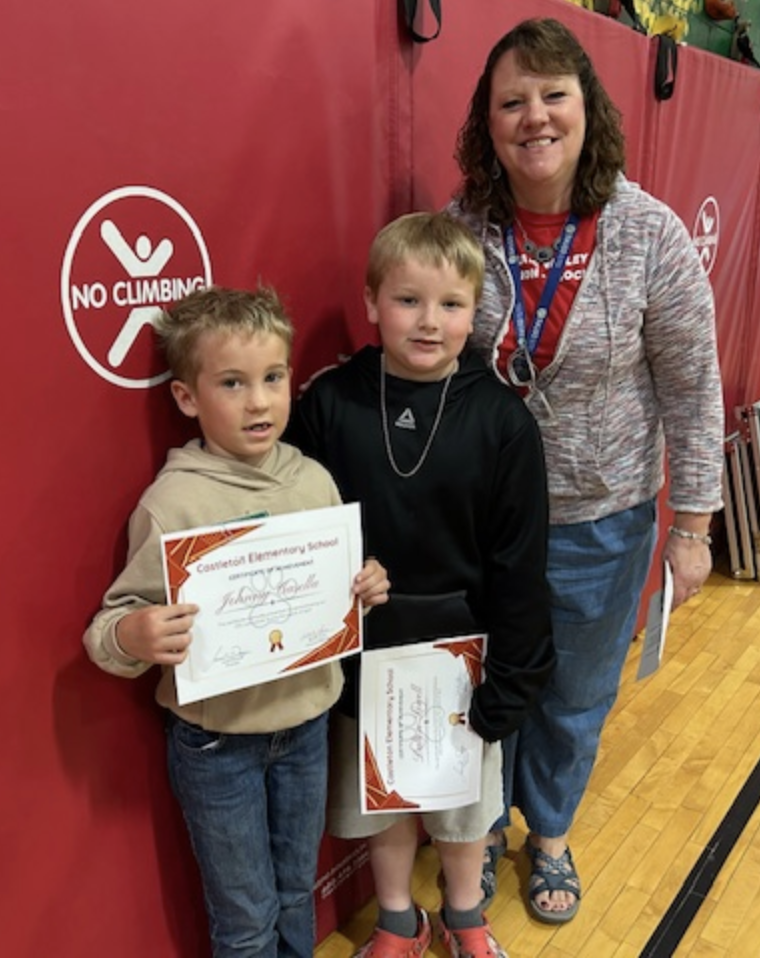 Two young students stand side by side holding certificates while a staff member stands behind them smiling. They are positioned in front of red gym wall mats with a visible “No Climbing” sign.