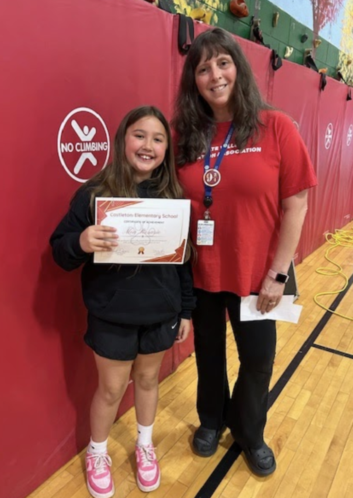 A student wearing a dark hoodie smiles while holding a certificate next to a staff member in a red shirt. They stand together in front of red gym wall mats.