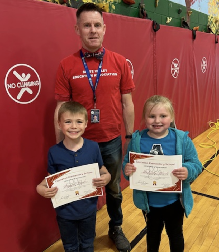 A staff member wearing a red shirt stands in a school gym beside two young students holding certificates from Castleton Elementary School. All three are smiling in front of red wall mats with a “No Climbing” sign.