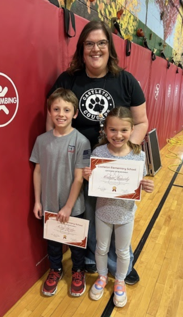 A staff member wearing a black shirt stands behind two smiling students holding certificates. They pose together in a school gym with red wall mats and gym equipment visible in the background.
