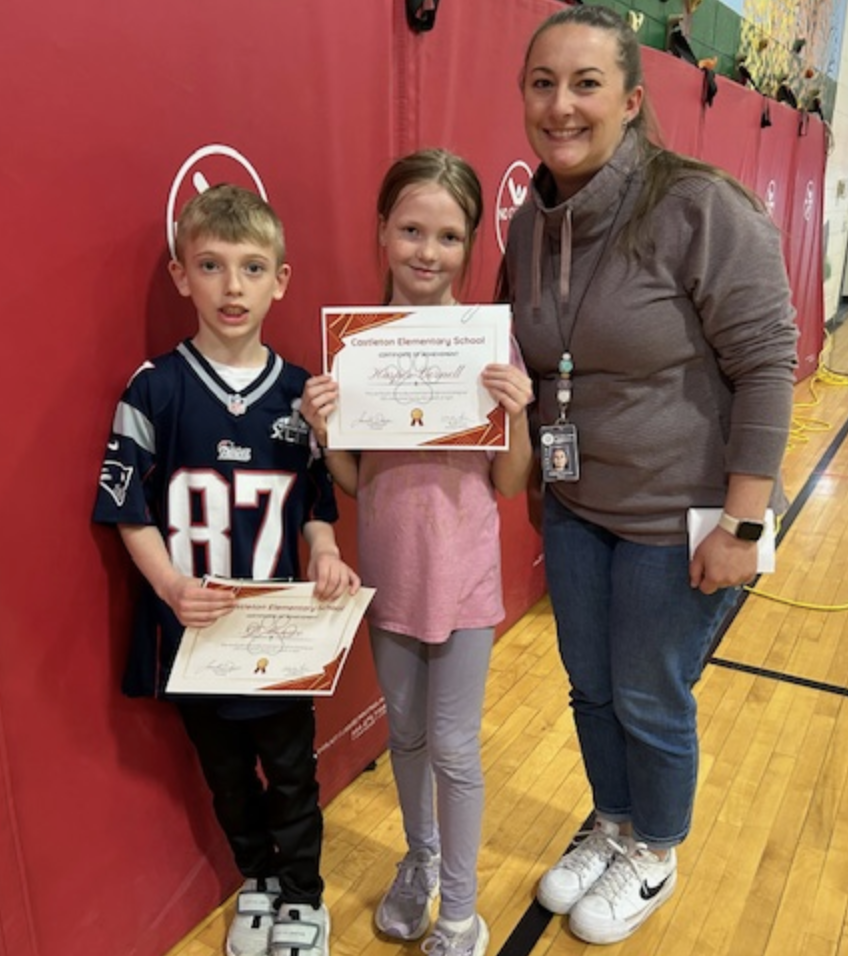 Two students hold up certificates while standing next to a smiling staff member. The group poses in a gymnasium with red padded walls and wooden flooring.