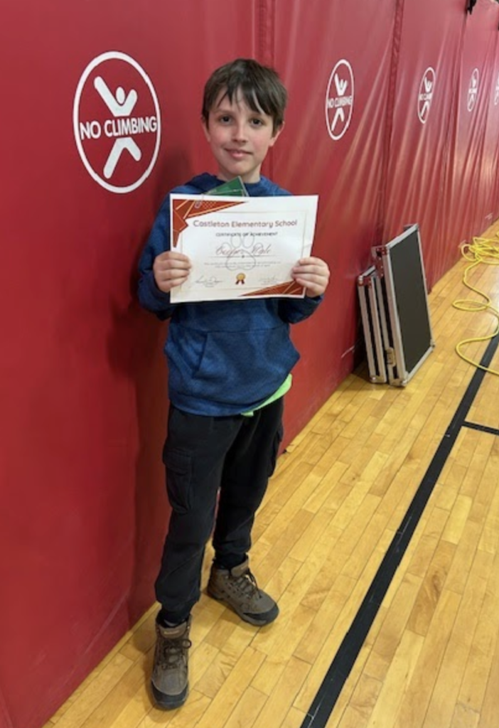 A single student stands holding a Castleton Elementary School certificate, smiling in a gymnasium with red wall padding and folded equipment nearby.