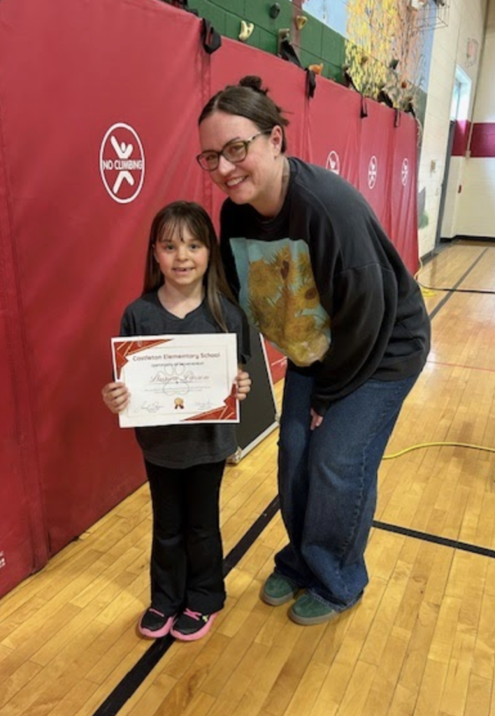 A staff member with glasses leans down next to a young student holding a certificate. Both are smiling in a school gym with red wall mats and gym flooring visible.