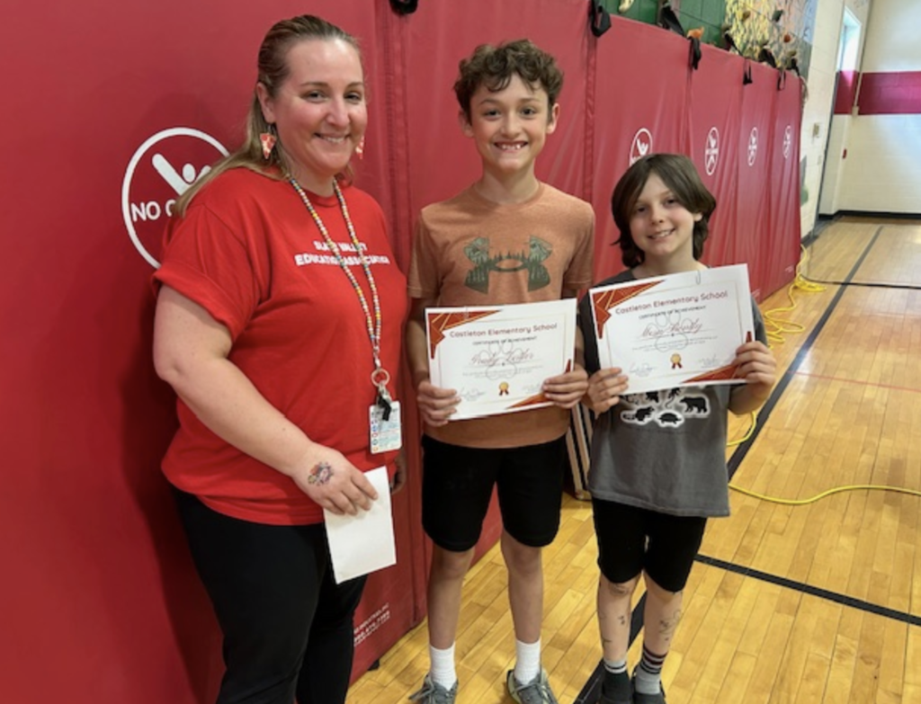 Two older elementary students hold certificates while standing beside a staff member in a red shirt. All three are smiling and posing in front of red gym wall mats.