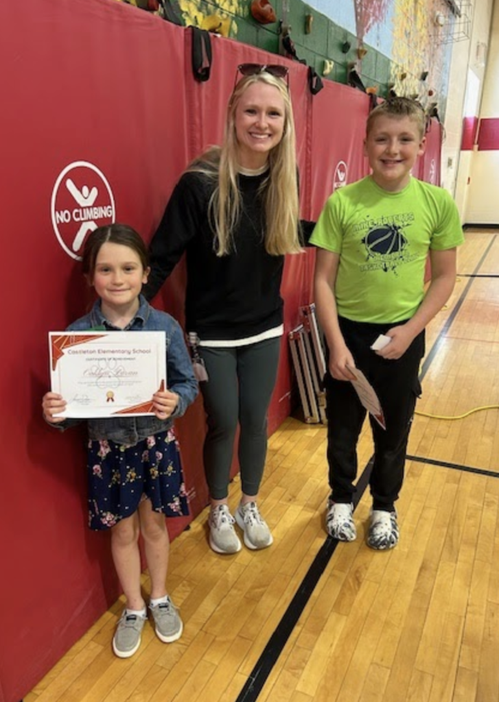 A staff member stands with two students—one holding a certificate—posing together in a gymnasium with red wall padding and equipment nearby.