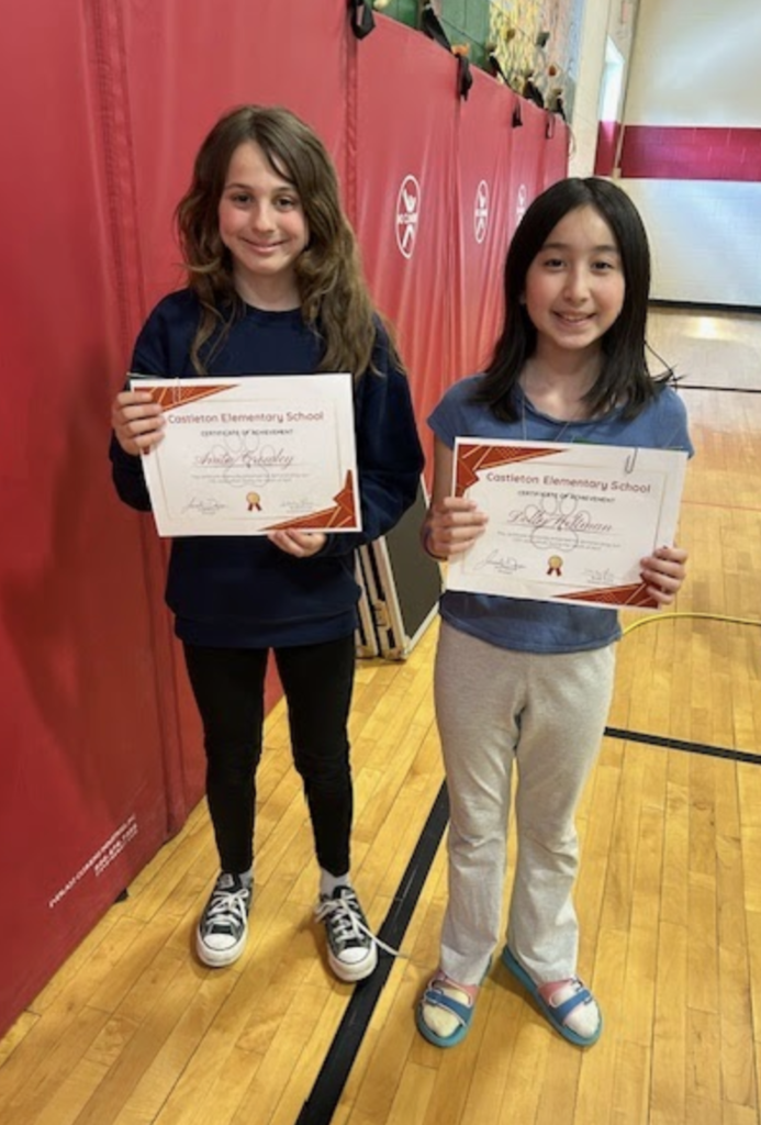 Two students stand side by side holding certificates and smiling in a school gym with red wall mats and folded equipment in the background.