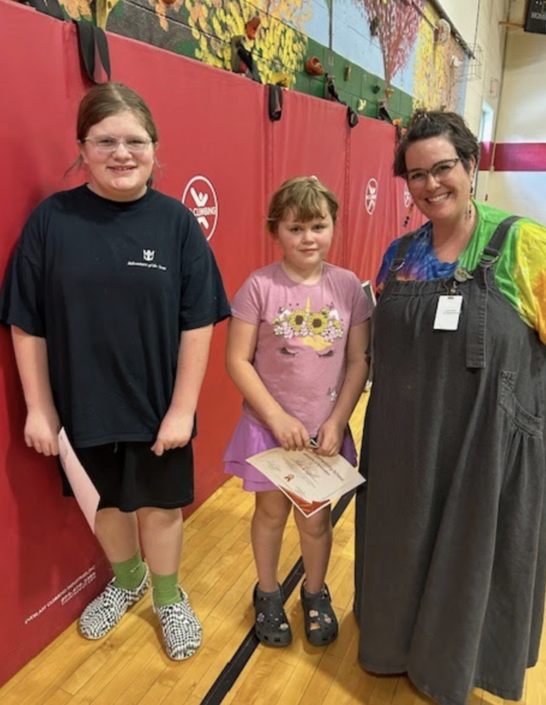 Two students stand with a staff member, smiling for a photo in a gym. One student holds certificates while the others hold papers.