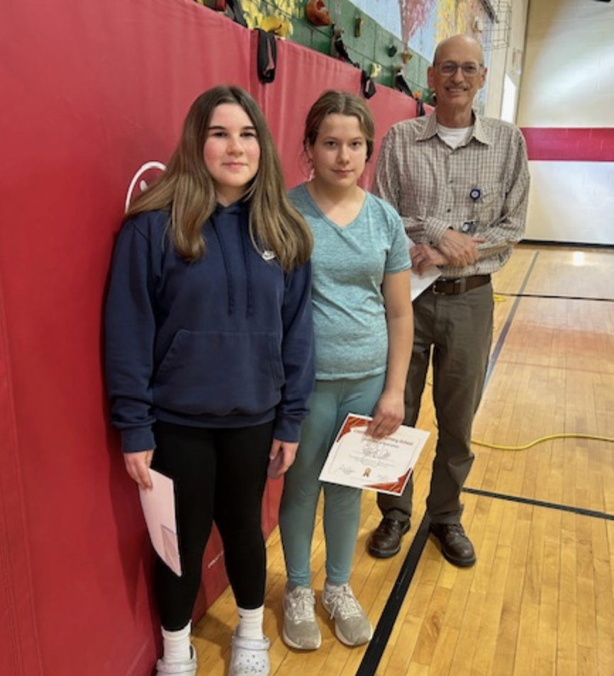 Two students stand side by side holding papers while a staff member stands slightly behind them smiling. They pose in a gym with red wall mats and wooden flooring.
