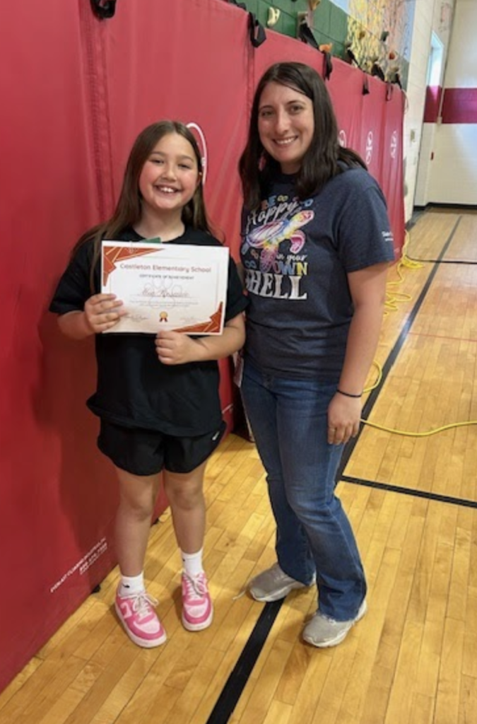 A smiling student holds a certificate next to a staff member. They stand together in front of red wall mats in a gymnasium.