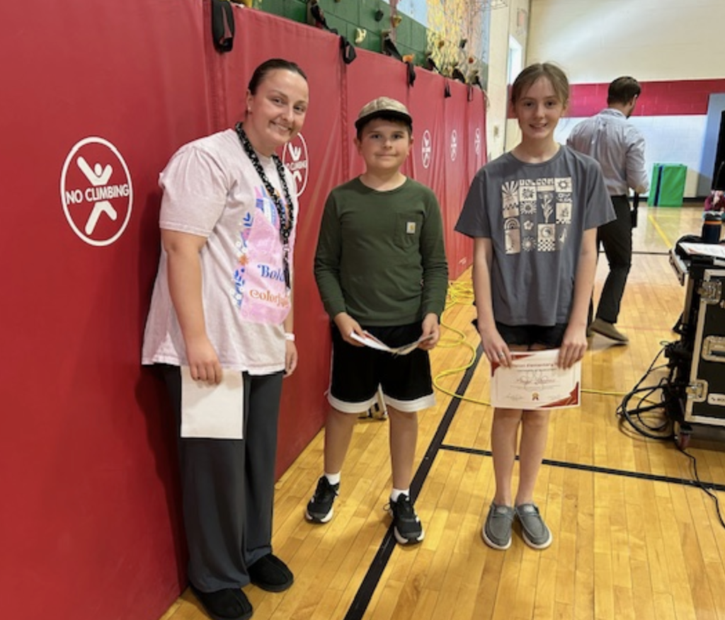 A staff member stands beside two upper elementary students holding certificates in a school gym. They smile in front of red wall mats with a “No Climbing” sign.