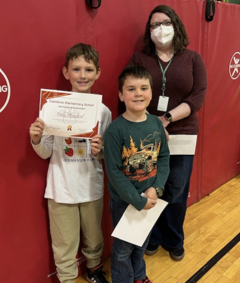 Two young students hold certificates while a staff member wearing a face mask stands behind them. They are positioned in front of red gym wall mats.