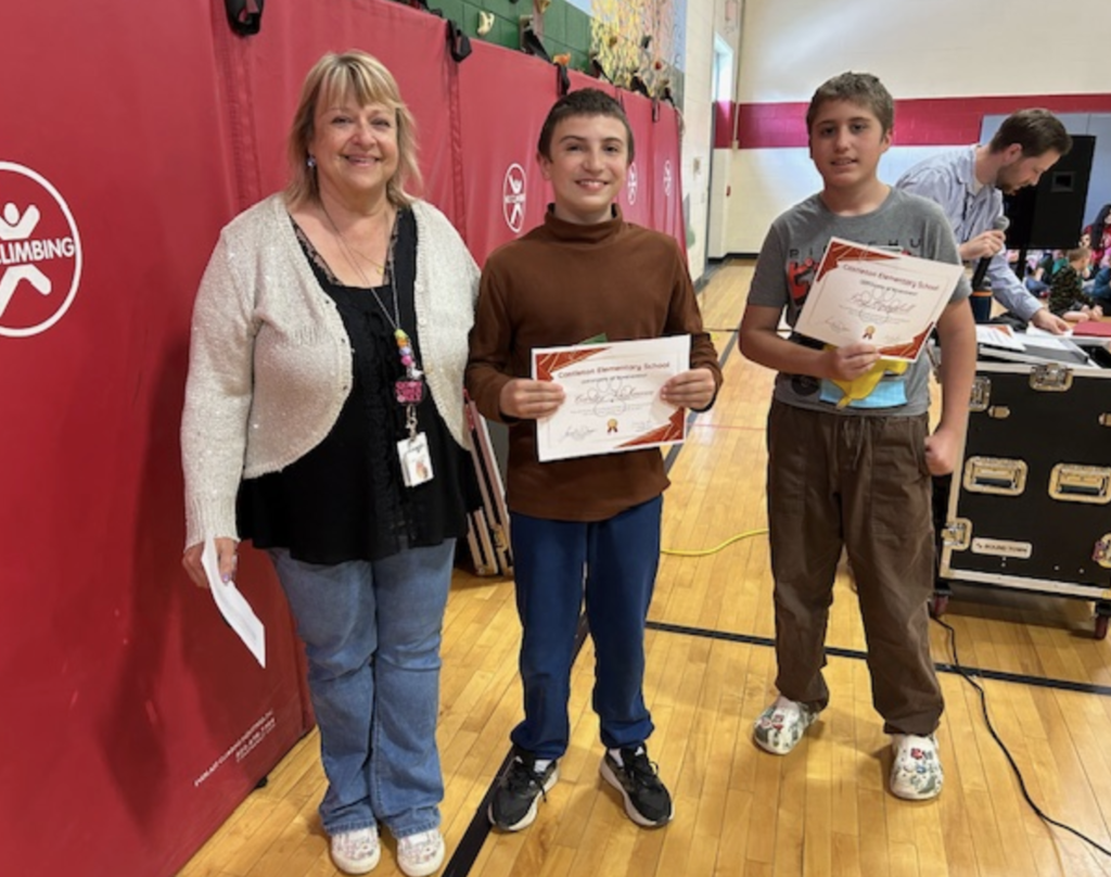 A smiling staff member stands with two students, each holding a Castleton Elementary School certificate. Gym equipment and red wall padding are visible behind them.