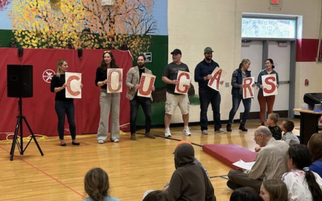 A group of parents stand in a gym holding large letters that spell “COUGARS” while students sit on the floor watching the presentation.