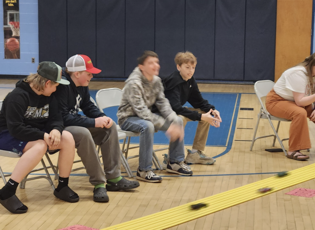 Several students sit along the side of a pinewood derby track in a gym as small cars race down the yellow track in front of them.