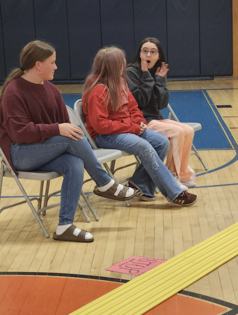 Three students sit in a row of folding chairs in a gym. One student reacts with surprise, hands on their face, while the others look on and smile.