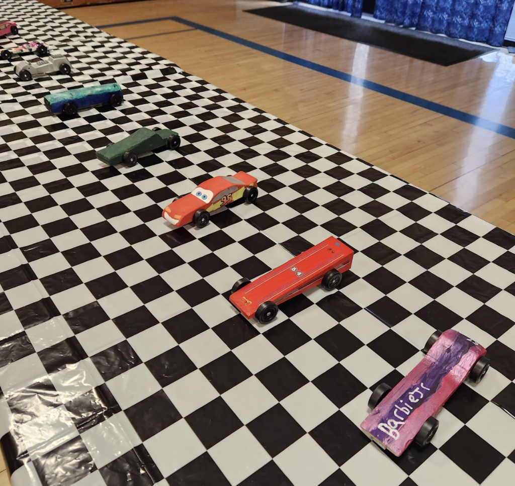 A row of handmade pinewood derby cars sits on a black-and-white checkered track in a school gym, including colorful designs like a red race car and a pink “Barbie” themed car.